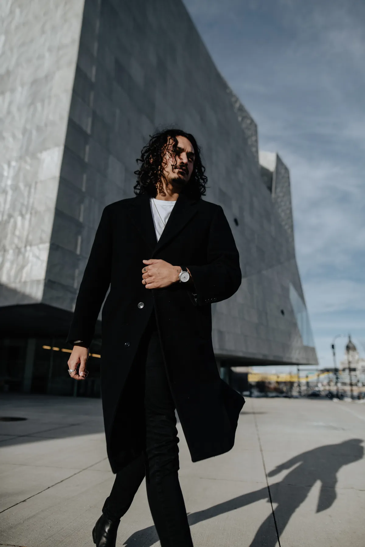 Walking past a concrete tower in a long black coat, a silver Aspect on black leather